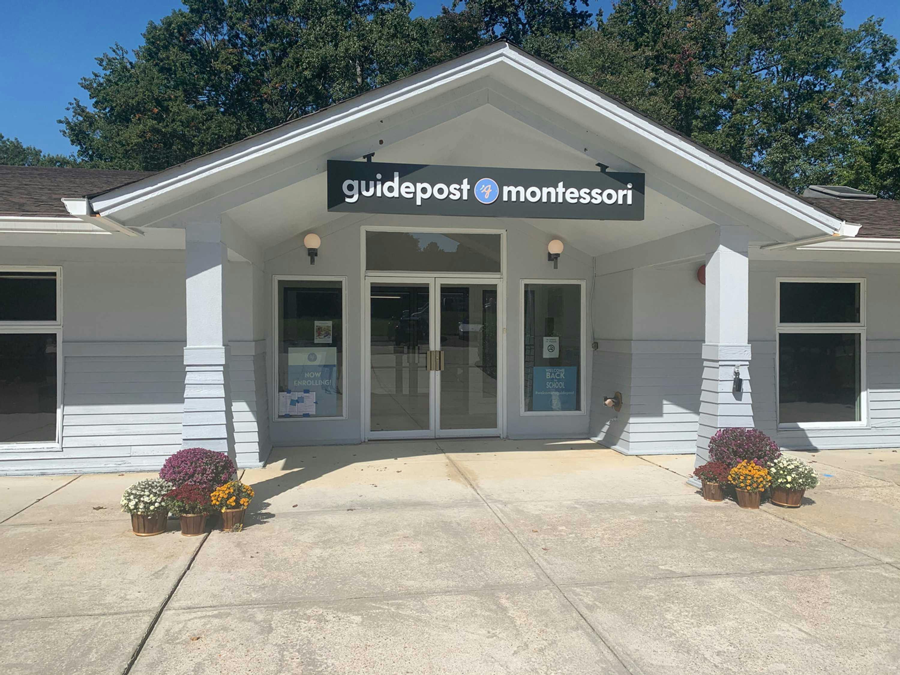 View of the entrance to Guidepost Montessori at Hampshire, showing the white exterior and front porch overhang with a branded Guidepost Montessori sign hanging from it. In front of both pillars to the overhang are flowers.