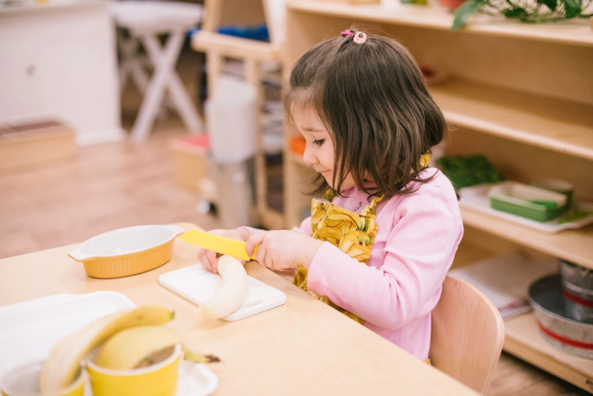 young girl sitting at a table in a Guidepost Montessori kindergarten classroom cutting a banana 