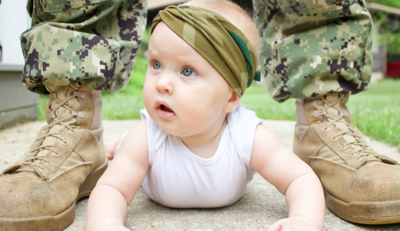 Infant with a military head wrap crawling on the ground in between the boots and legs of a military serviceman. 