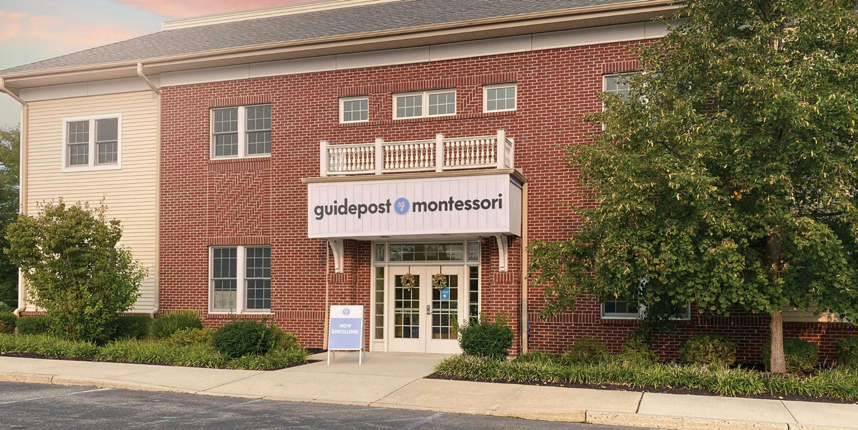 View of Guidepost Montessori at Somerset from the parking lot that shows the two story building with a brick exterior. A fake balcony rests above the entrance door and a branded Guidepost Montessori sign hangs form it. Shrubs and bushes line the building.