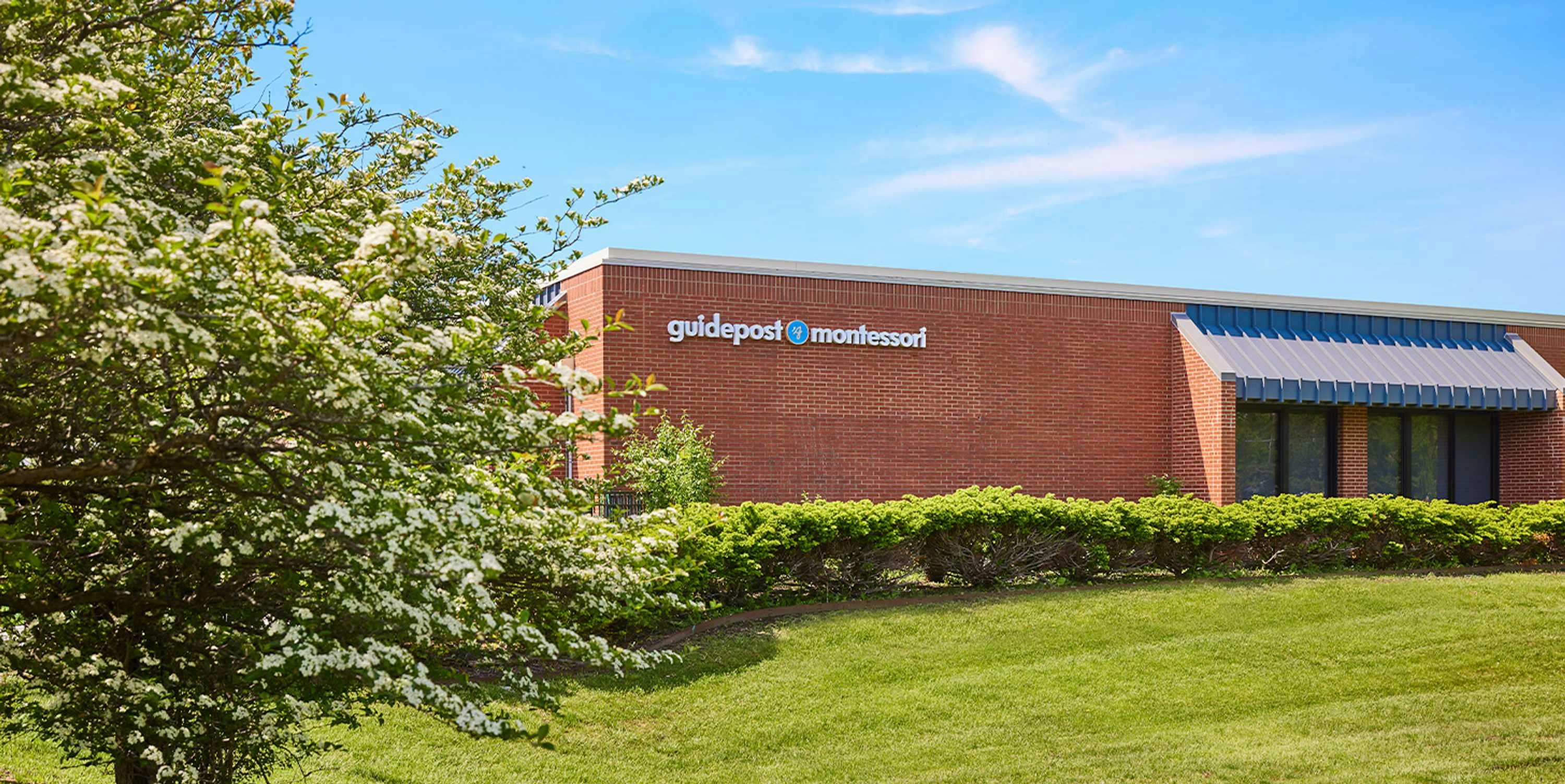 View of Guidepost Montessori at Deerbrook from a sprawling green lawn, showing the square, brick building with a branded Guidepost Montessori sign as well as a connected structural awning over some windows.