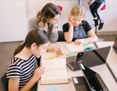 A teacher helps students learn how to read in a classroom setting, with sticky notes, open laptops and a student on a barstool in the background. 