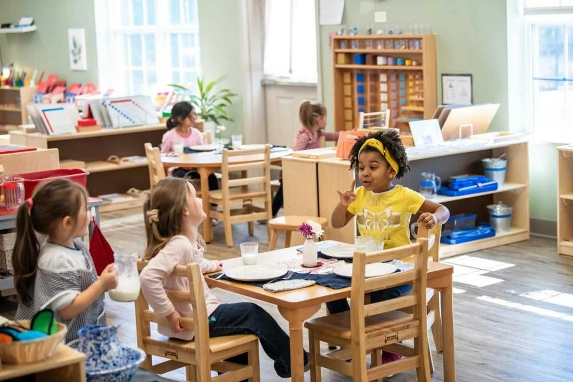 Three kindergarten-aged school girls enjoy a conversation over glasses of milk, while two other students complete activities behind them in the well-organized Montessori classroom. 