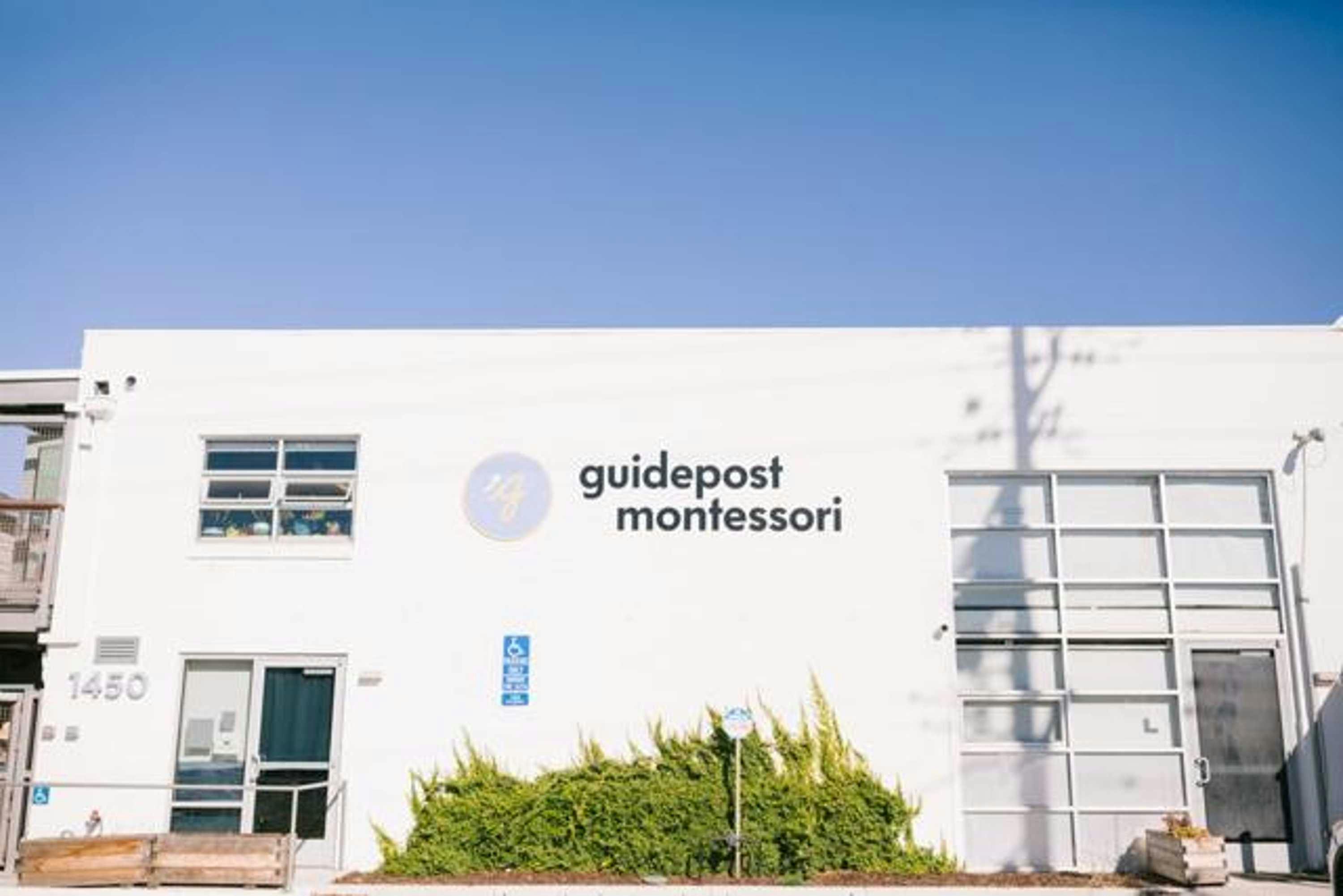 View of Guidepost Montessori at Emeryville facade with a large Guidepost Montessori branded sign which hangs above a bush and a street sign.
