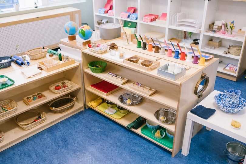 Shelves are stacked with colorful paper and organized with classroom tools like scissors, pens, paintbrushes and glue. Students complete work in the background.