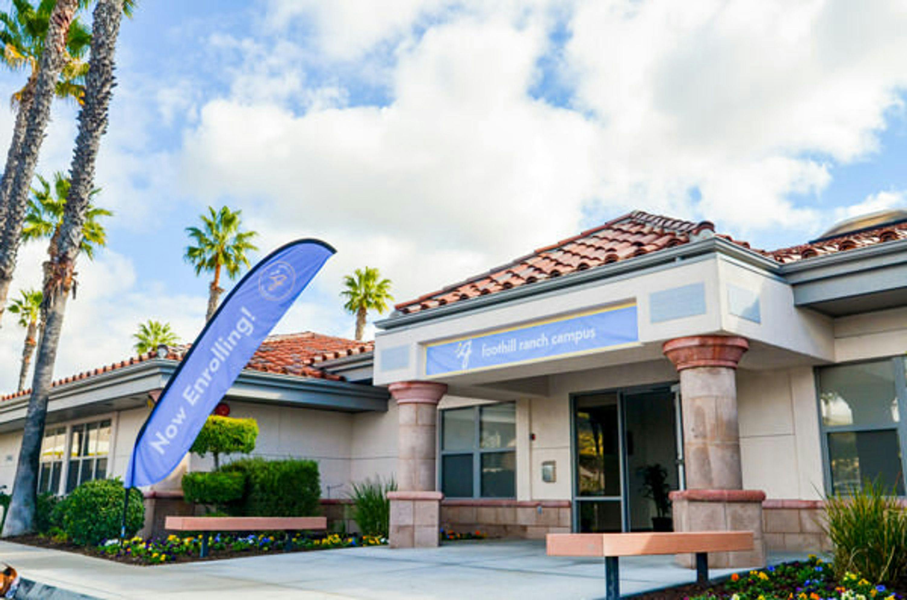 View of Guidepost Montessori at Foothill Ranch entrance way with sitting benches on either side, colorful flower gardens, palm trees towering in the background and a feather "Now Enrolling" Banner.