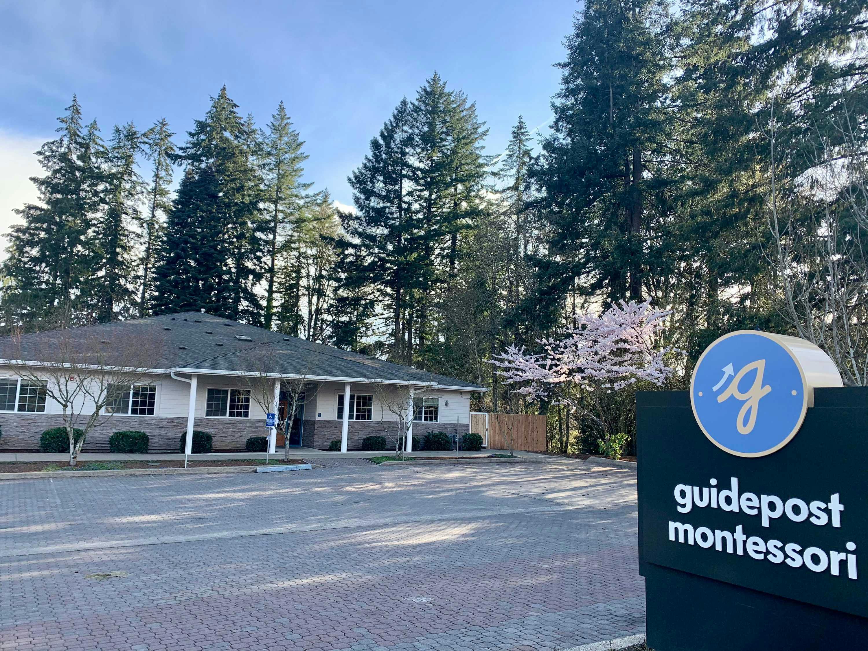 View of Guidepost Montessori at South Beaverton that shows the brick inlaid parking lot and a street-facing branded Guidepost Montessori sign. There are a lot of tall trees in the background, some smaller trees in the parking lot and one of them is blossoming pink flowers.